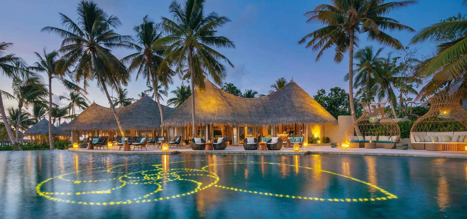 Naiboli's poolside pavilion lit at dusk, palm trees silhouetted against the pool's floating flower display.