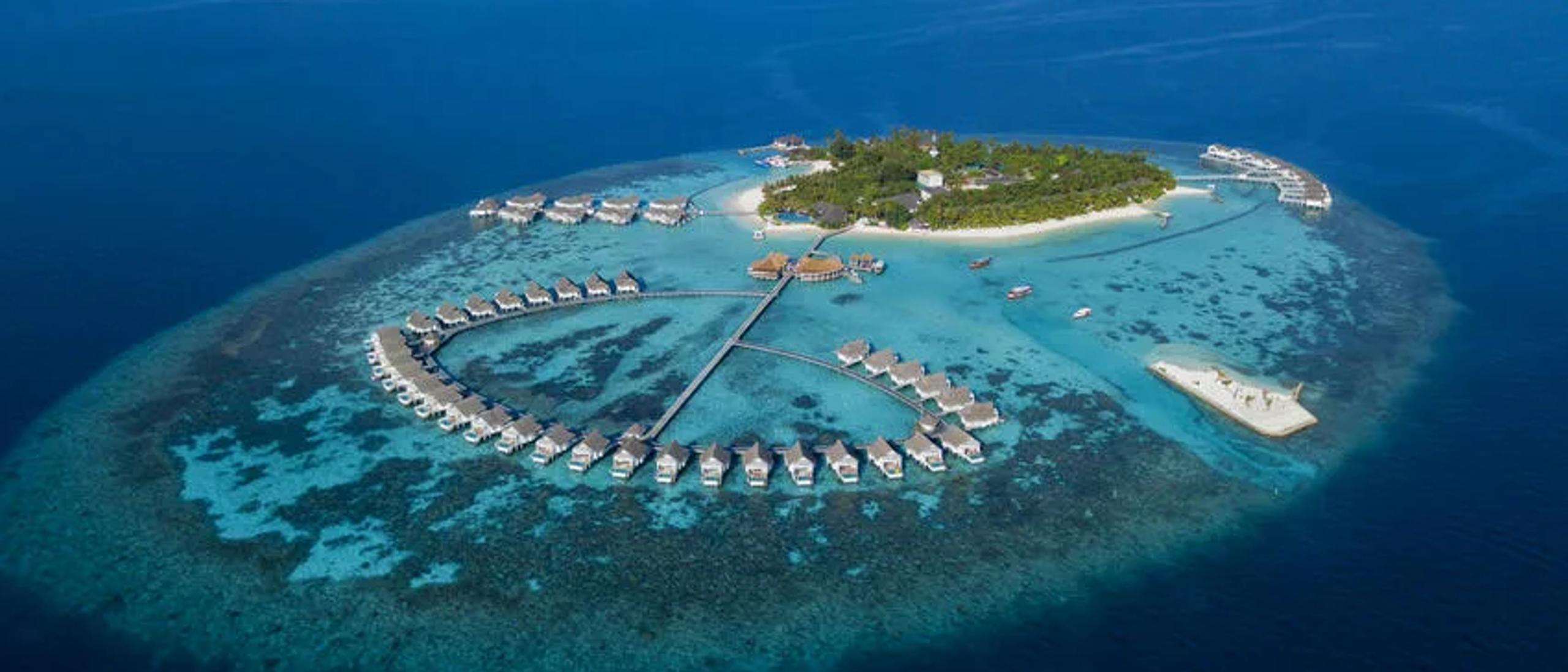 Aerial view of the full island, overwater villas ringing the reef edge with open ocean beyond