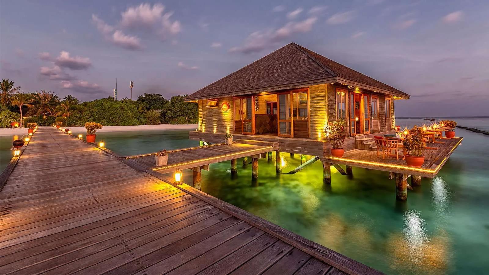 Aqua Restaurant lit up at dusk, its overwater dining room reached by a long timber jetty lined with lanterns