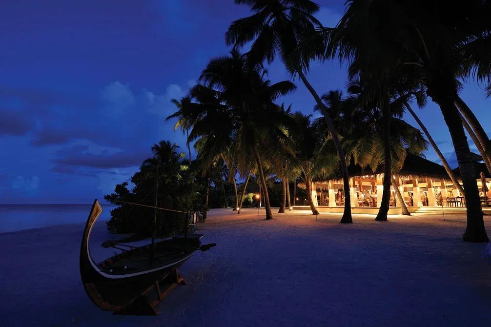 Magu's beachfront dining room glowing at dusk, a traditional dhoni resting on the sand in front