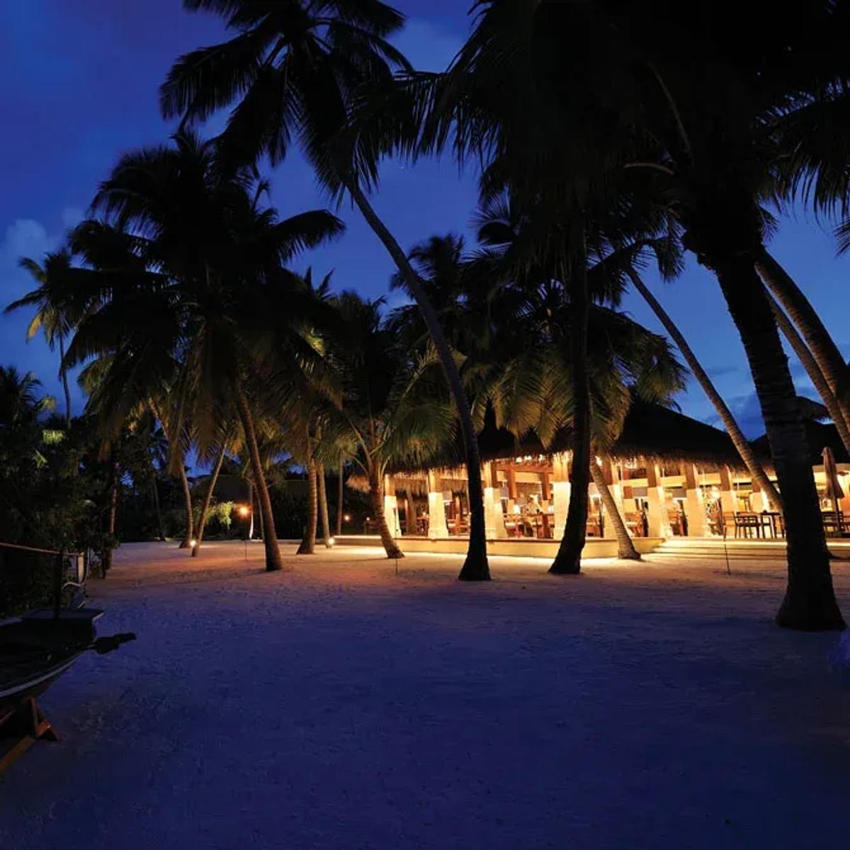 Magu's beachfront dining room glowing at dusk, a traditional dhoni resting on the sand in front