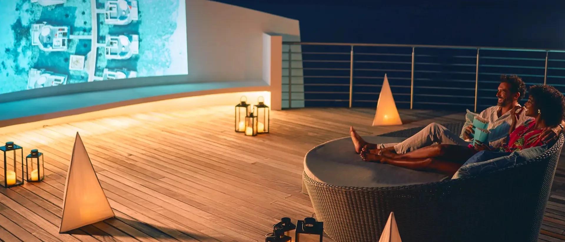 A couple relaxing on a rooftop daybed at night, watching a film projected onto an outdoor screen