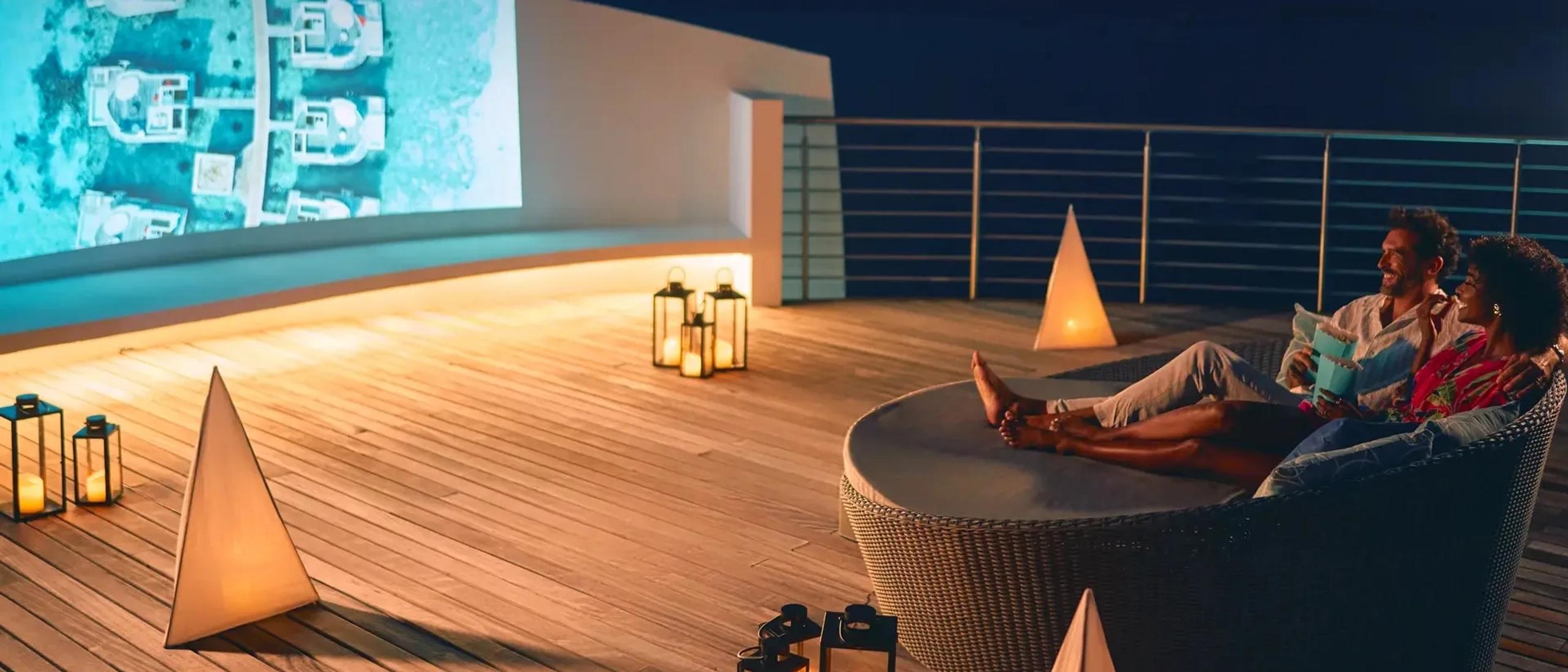 A couple relaxing on a rooftop daybed at night, watching a film projected onto an outdoor screen, lanterns lit around them