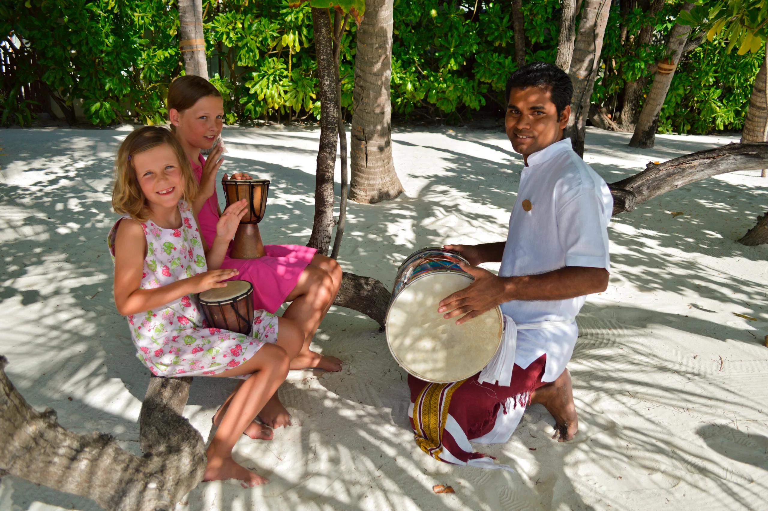 Two children playing hand drums with a Coconut Tribe staff member on the beach, shaded by trees