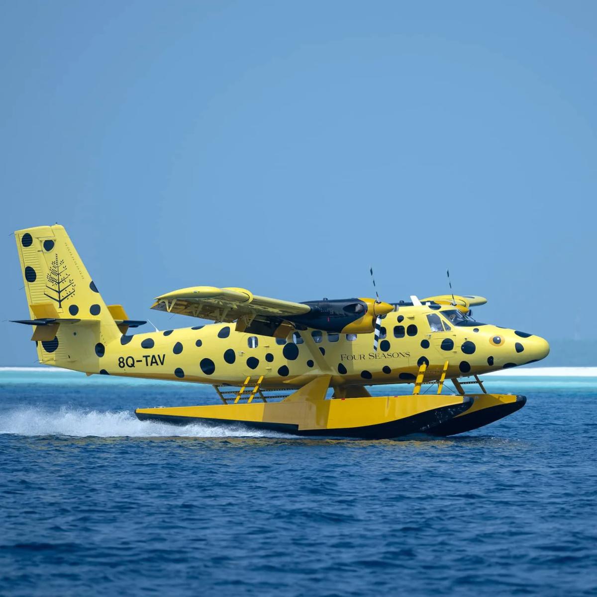 A yellow spotted Four Seasons seaplane skimming across the lagoon at speed, spray fanning out behind it and the reef line visible in the distance