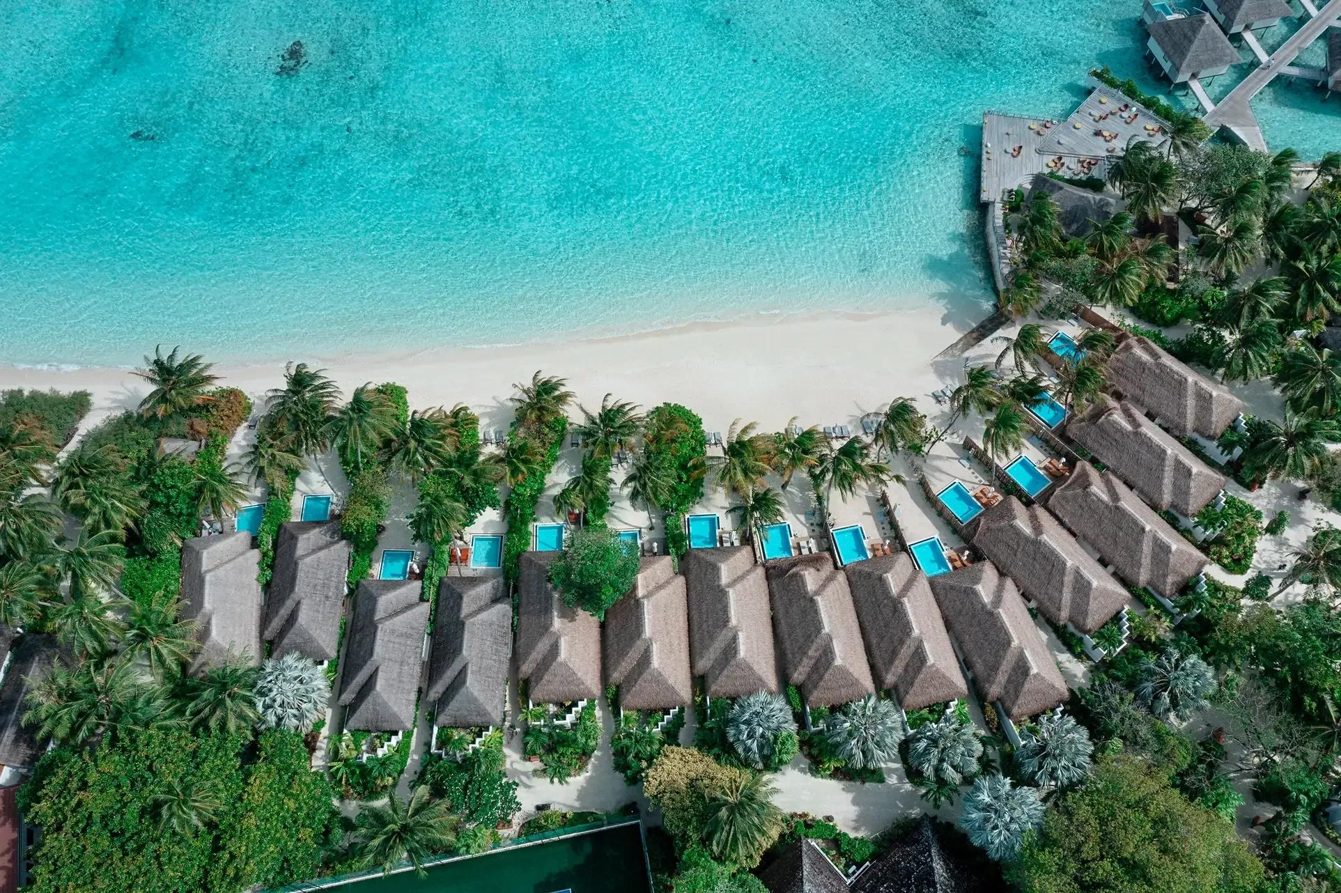 An aerial view of Velassaru's Beach Villas with Pool lined along the shore, overwater villas visible beyond across the lagoon
