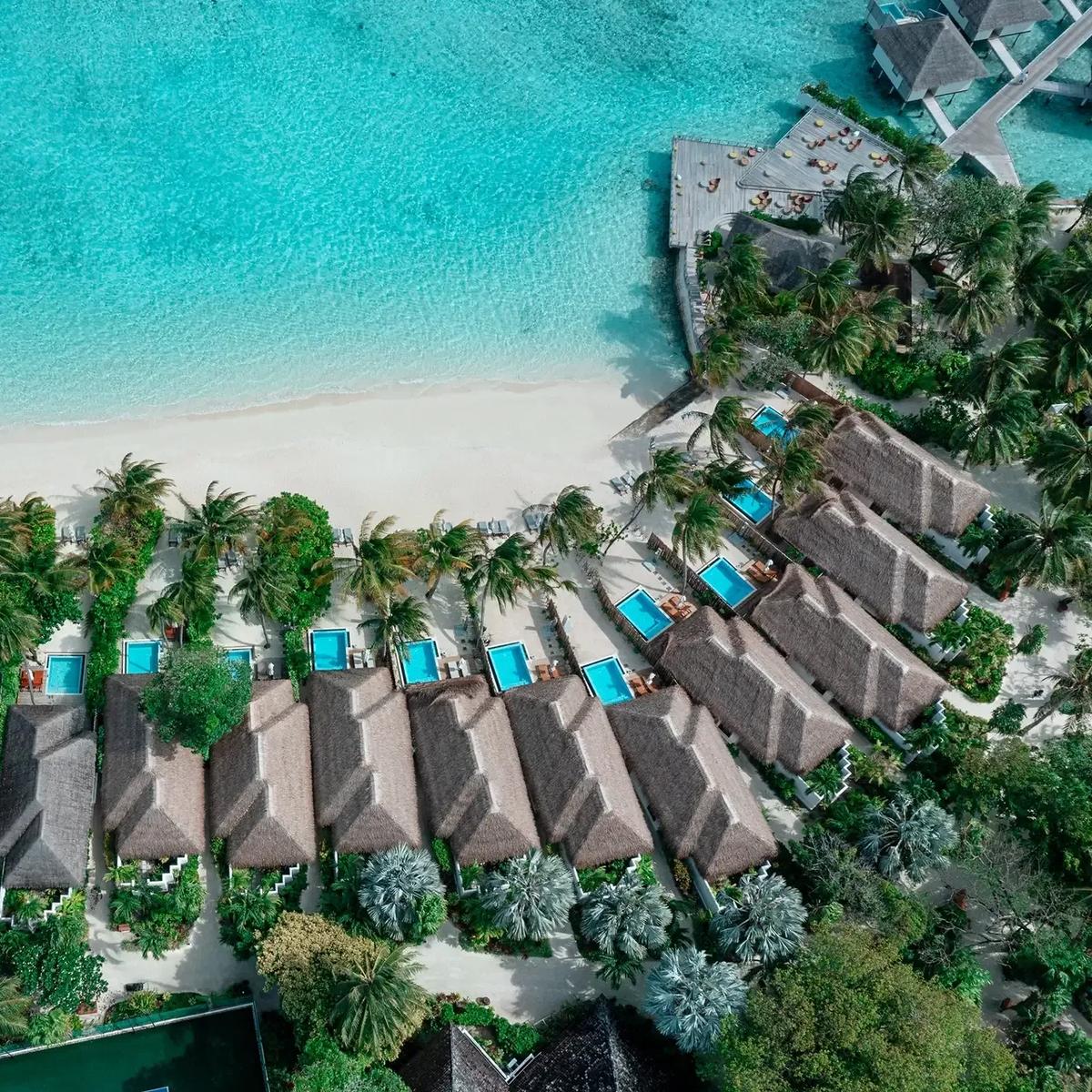 An aerial view of Velassaru's Beach Villas with Pool lined along the shore, overwater villas visible beyond across the lagoon