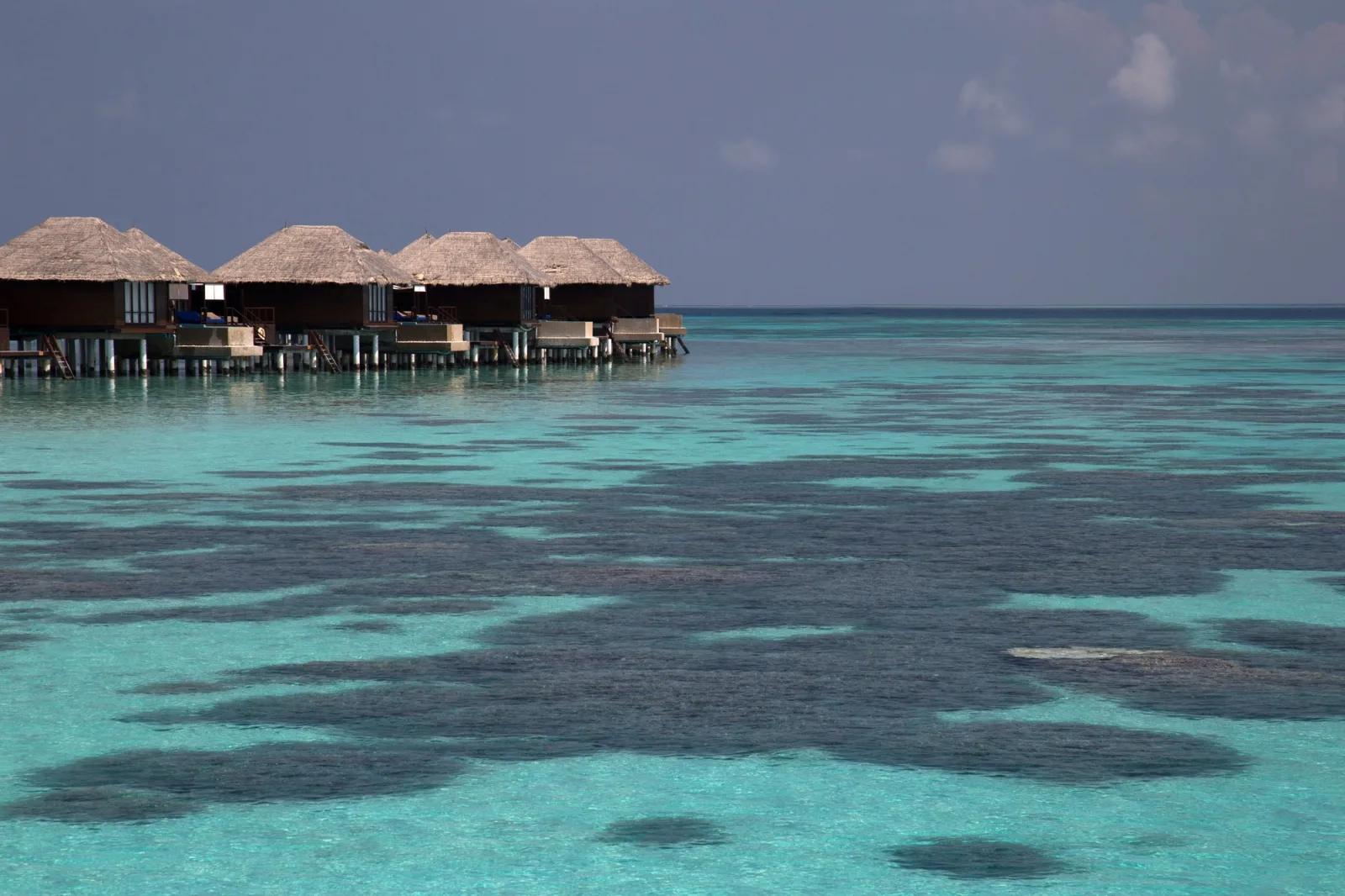 A row of thatched-roof overwater bungalows at dusk, turquoise water and dark reef patches below
