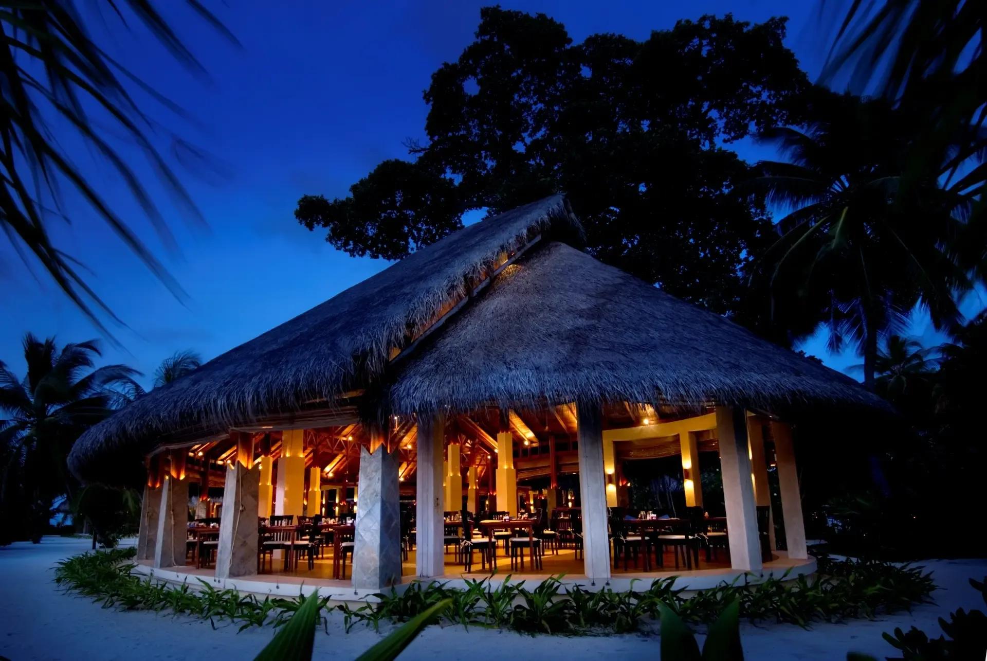 The thatched triple-peaked roof of the Malaafaiy, Haruge and Farivalhu buffet restaurant, lit from within at dusk beneath large canopy trees