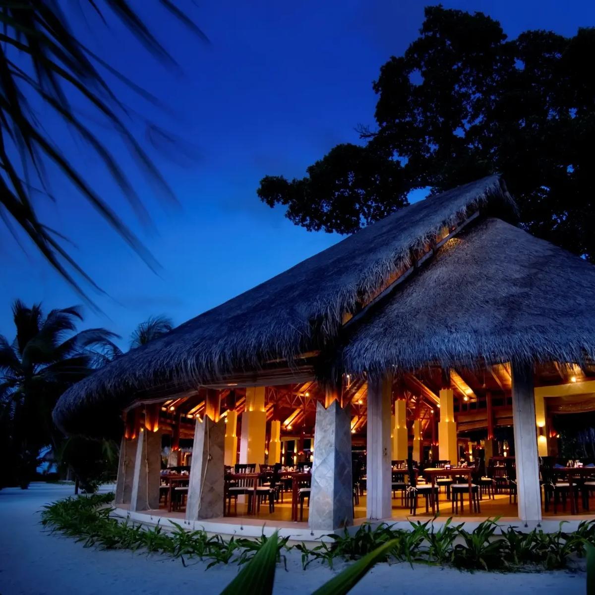 The thatched triple-peaked roof of the Malaafaiy, Haruge and Farivalhu buffet restaurant, lit from within at dusk beneath large canopy trees