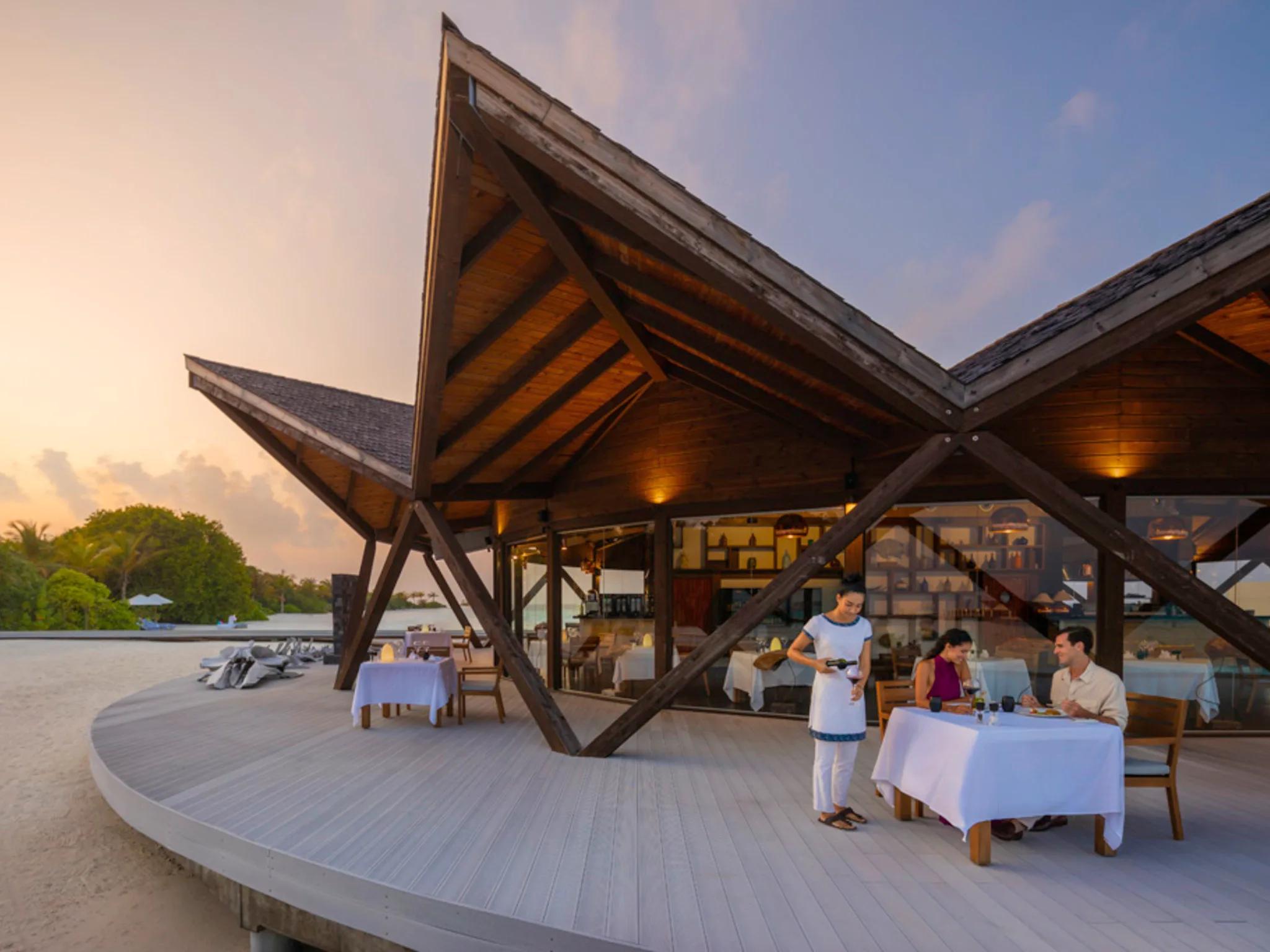 A waiter pouring wine for a couple dining at dusk beneath Bodumas' angular wood-beamed roof, tables set along a curved deck on the beach