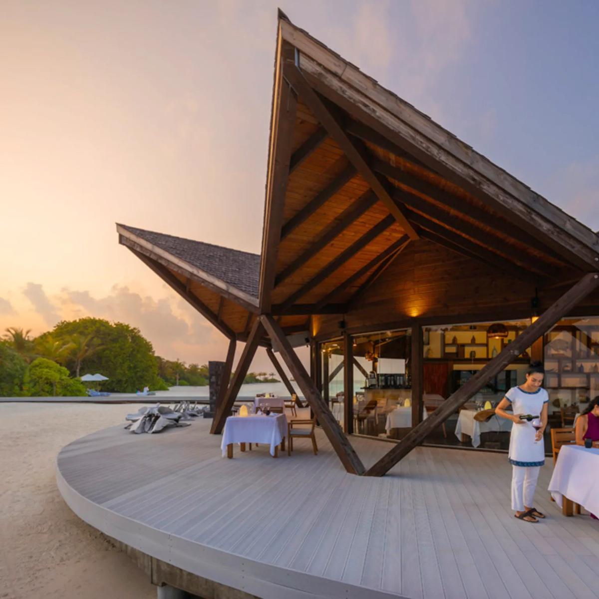 A waiter pouring wine for a couple dining at dusk beneath Bodumas' angular wood-beamed roof, tables set along a curved deck on the beach