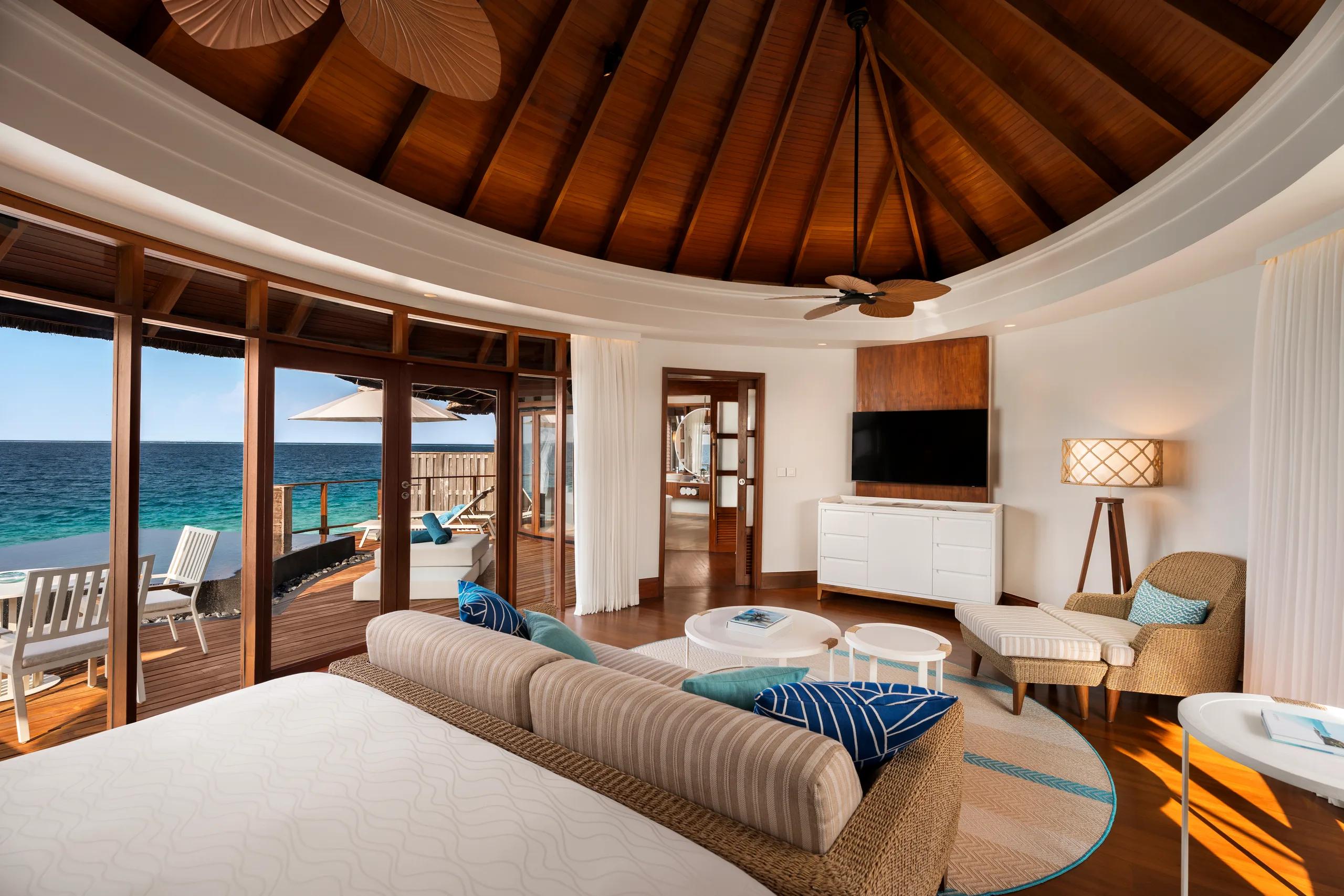 The interior of a Water Villa looking out through open doors to a shaded deck and the ocean beyond, thatched dome ceiling overhead