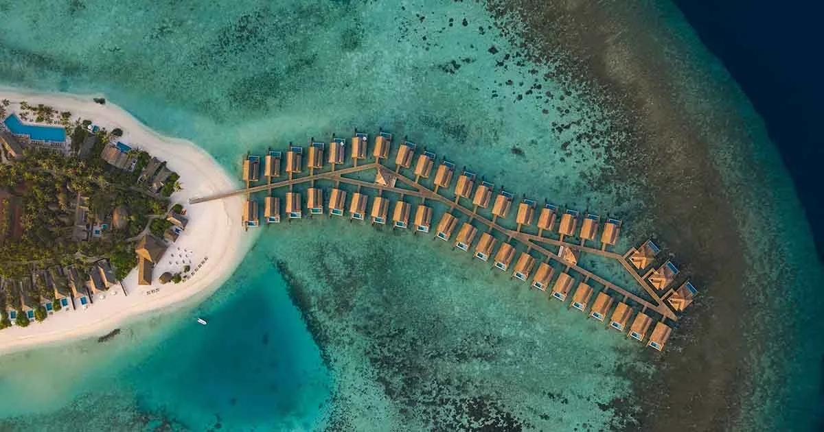 Emerald Faarufushi's island and lagoon seen from above, overwater villas on one jetty and the beach curving beside them