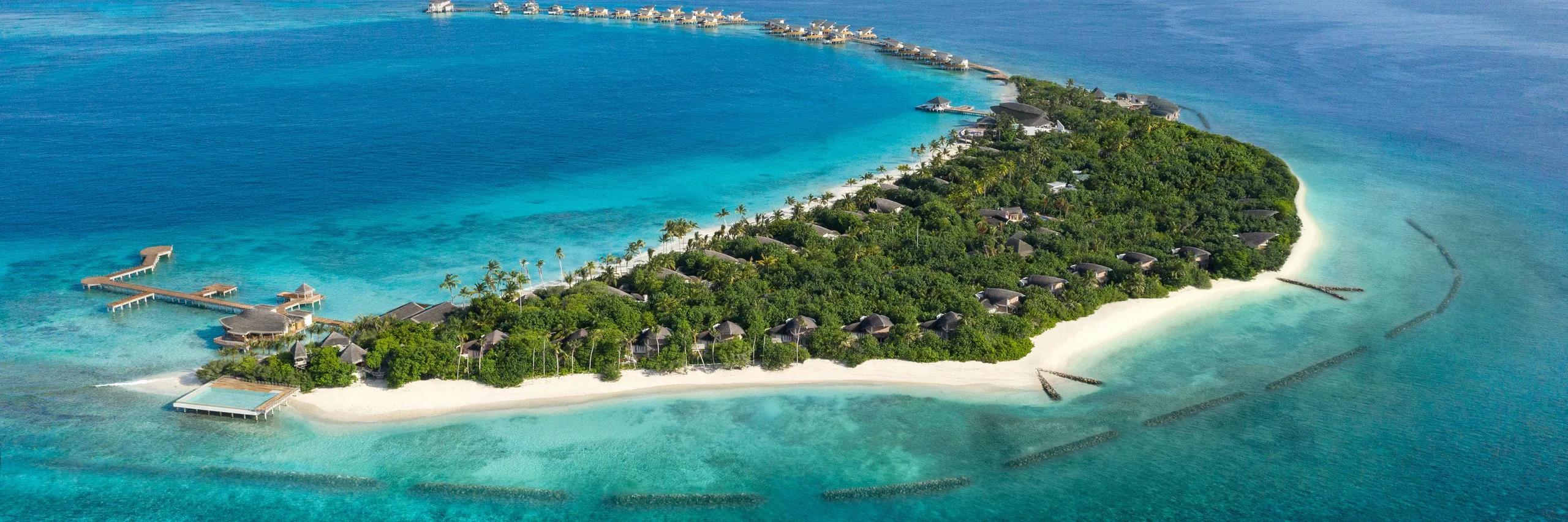 Aerial view of Vagaru Island, JW Marriott Maldives, showing beach villas and jungle canopy tapering to a row of overwater villas on stilts, ringed by the lagoon