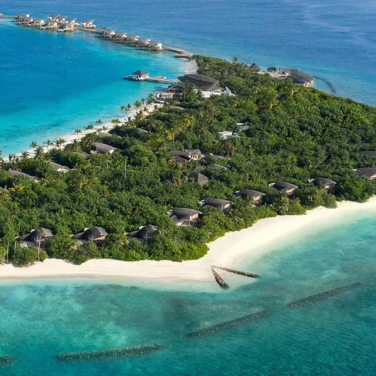 Aerial view of Vagaru Island, JW Marriott Maldives, showing beach villas and jungle canopy tapering to a row of overwater villas on stilts, ringed by the lagoon