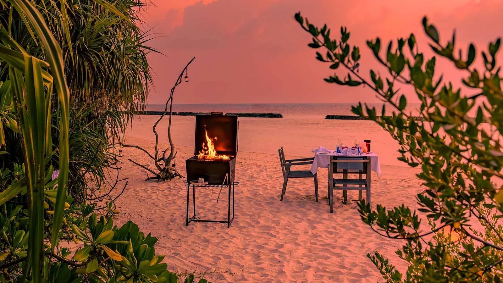 A private beach dinner set up at dusk, a lit barbecue beside a single table laid on the sand
