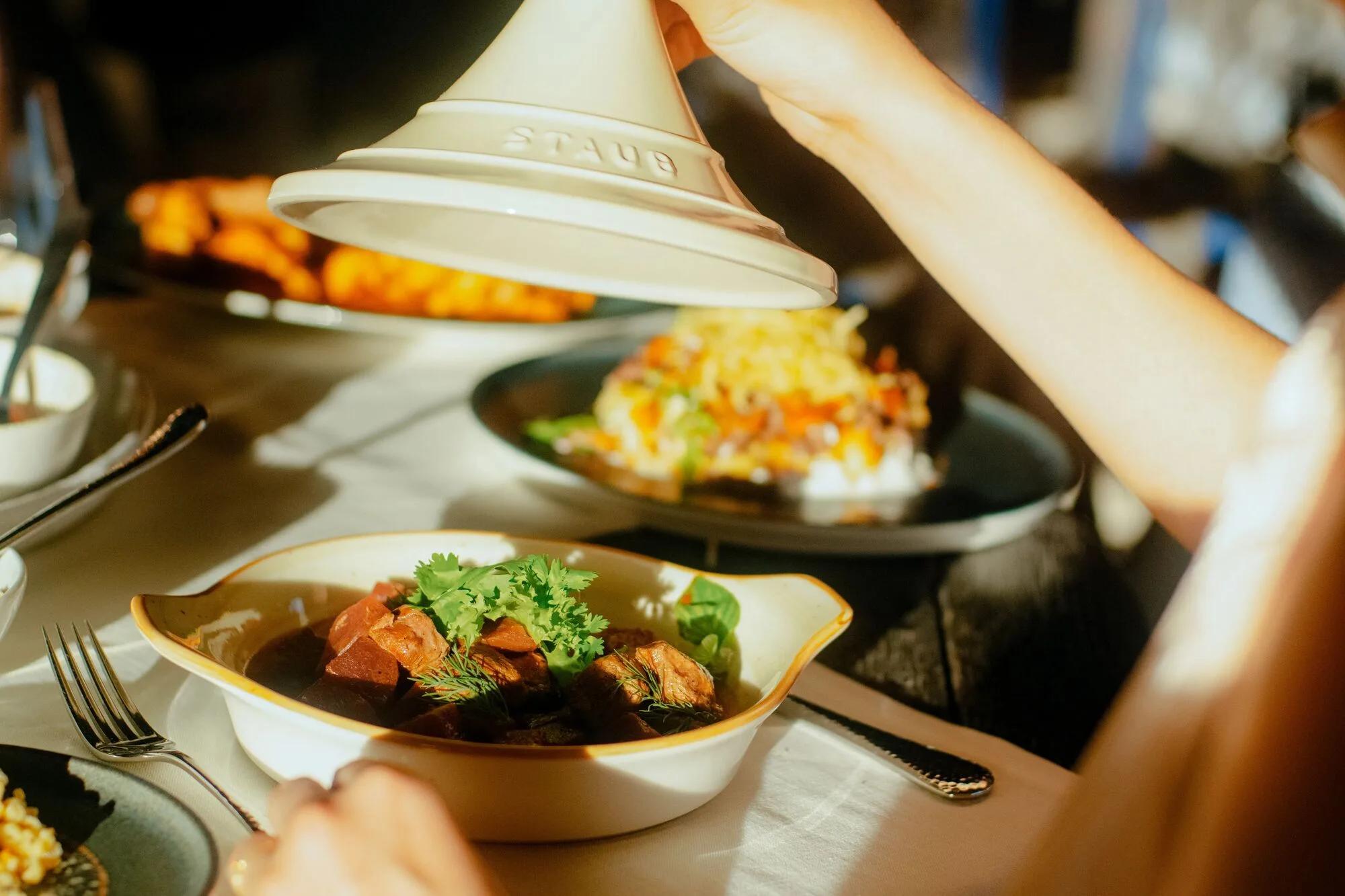A waiter lifting the lid off a lamb and beet tagine at TUH'U, coriander and herbs on top