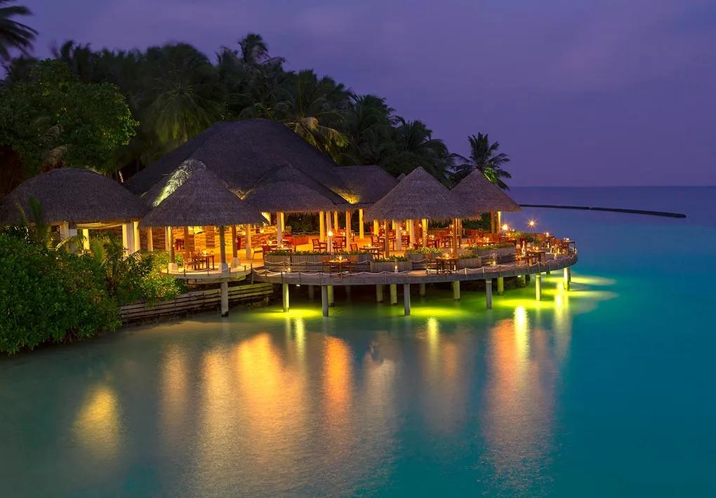 Cayenne Grill's thatched-roof dining pavilions lit at dusk, connected by a jetty reaching out over the lagoon