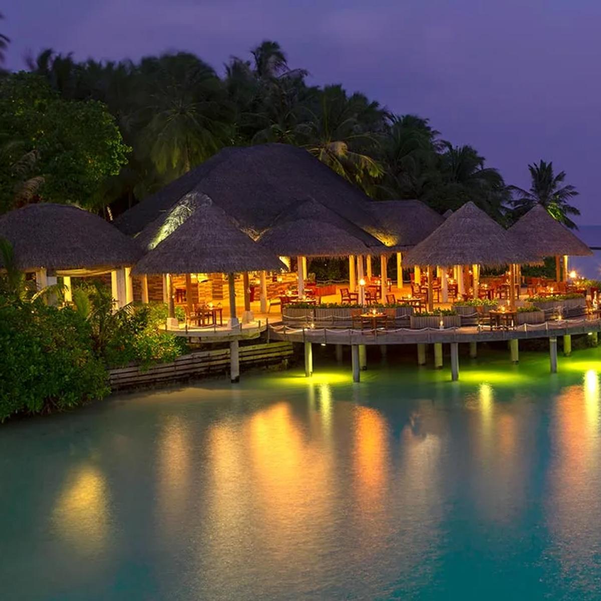 Cayenne Grill's thatched-roof dining pavilions lit at dusk, connected by a jetty reaching out over the lagoon
