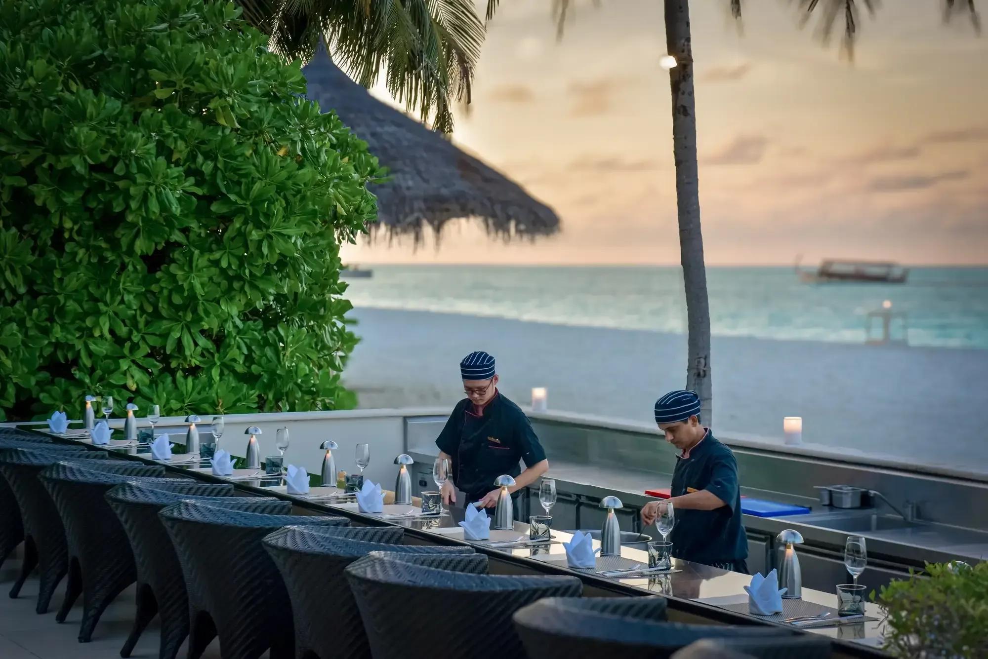 Chefs preparing the teppanyaki grill at Koko Grill at sunset, palm trees and the beach behind them at Conrad Maldives Rangali