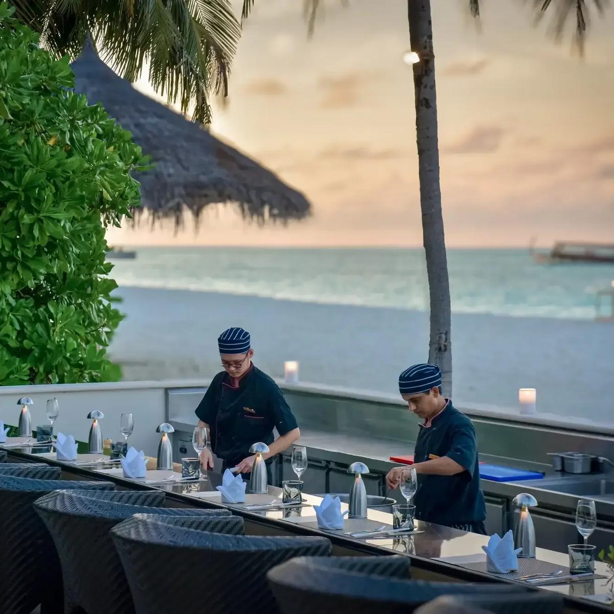 Chefs preparing the teppanyaki grill at Koko Grill at sunset, palm trees and the beach behind them at Conrad Maldives Rangali