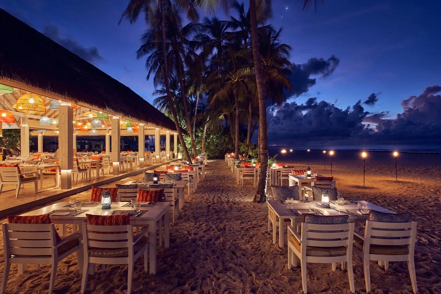 Beach Kitchen's open-air dining pavilion strung with colourful pendant lights, tables set beneath a thatched roof opening onto the sand at dusk
