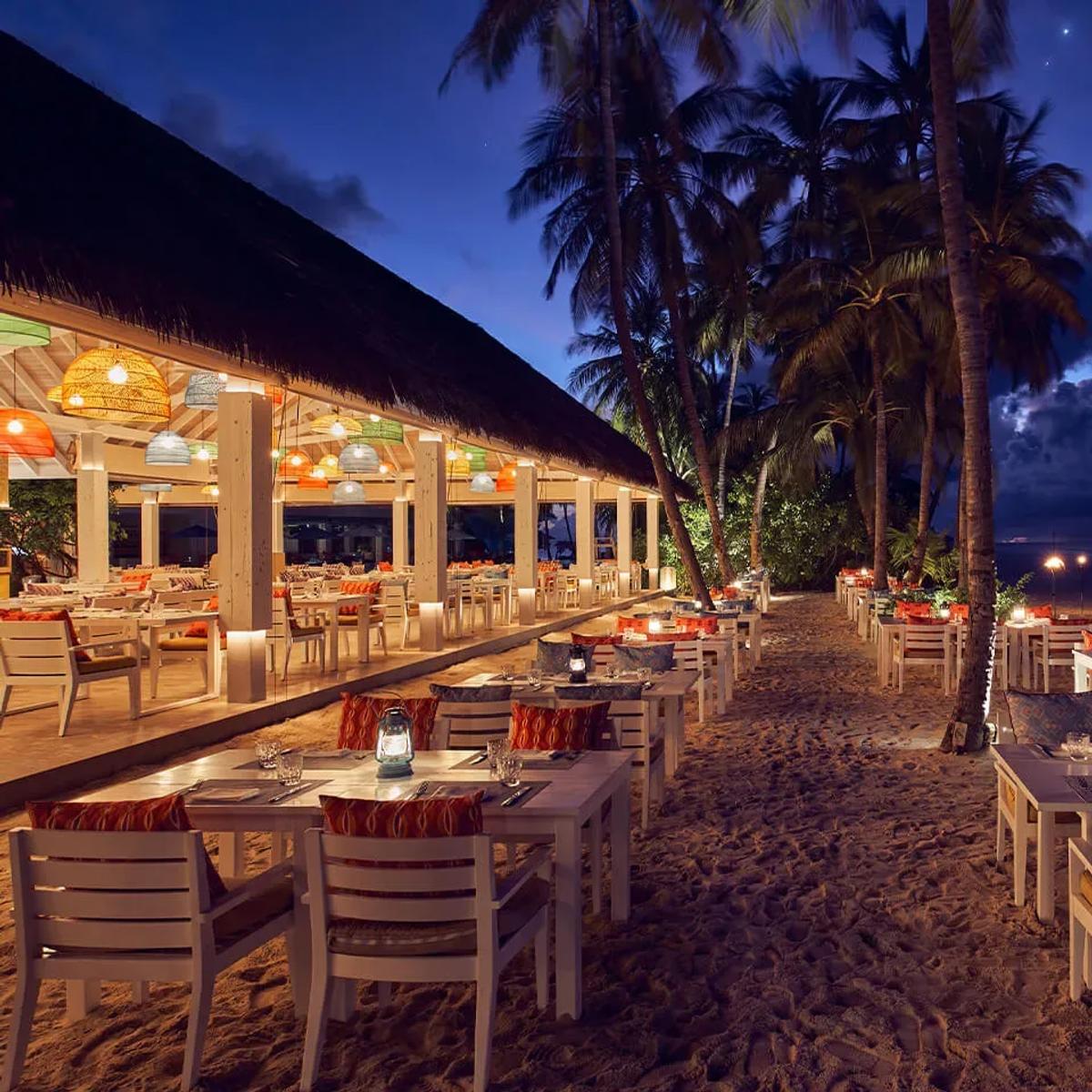 Beach Kitchen's open-air dining pavilion strung with colourful pendant lights, tables set beneath a thatched roof opening onto the sand at dusk