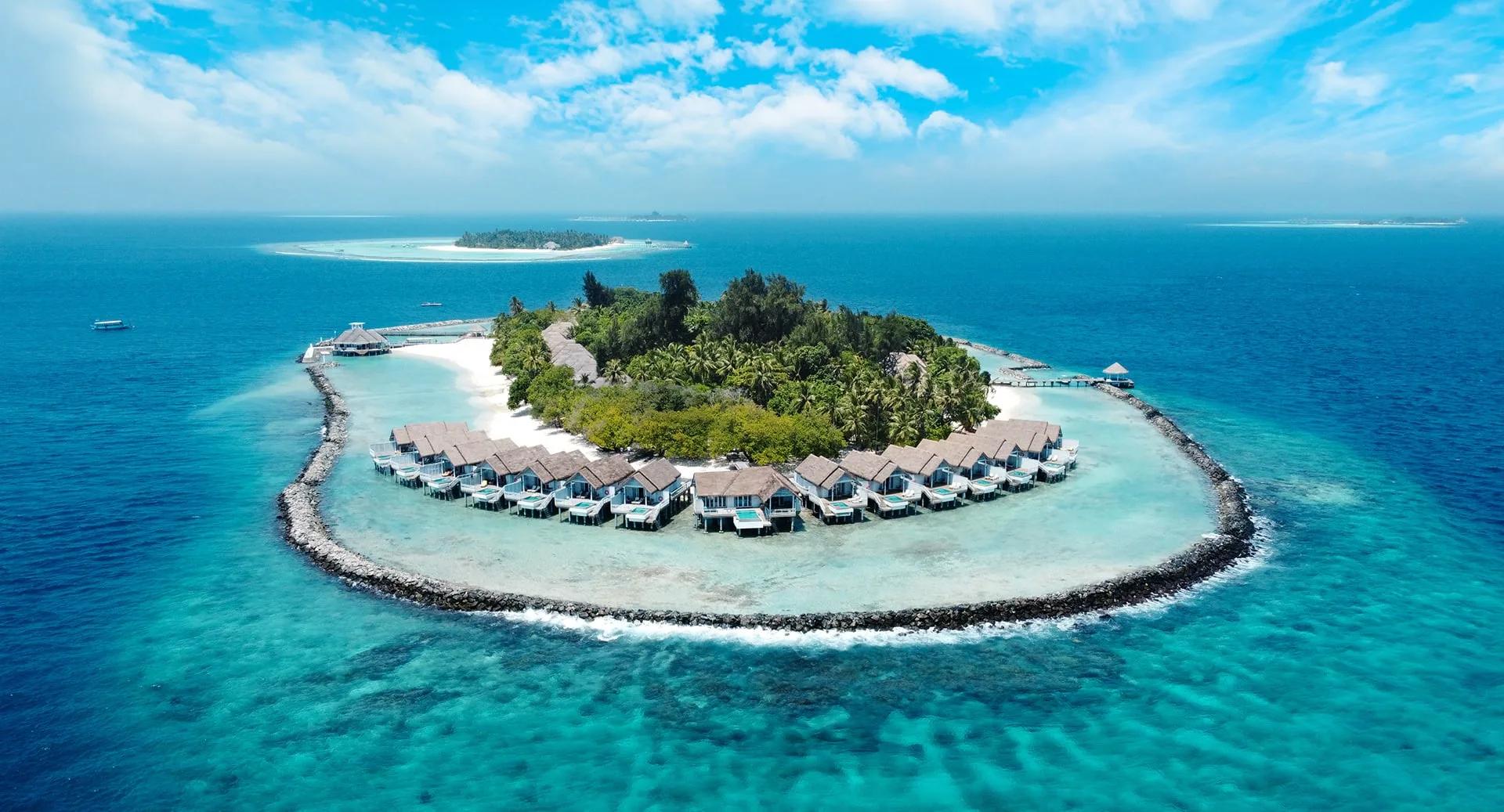 An aerial view of NH Maldives Kuda Rah Resort's small circular reef island, ringed by a rock breakwater with overwater villas stepping into the lagoon