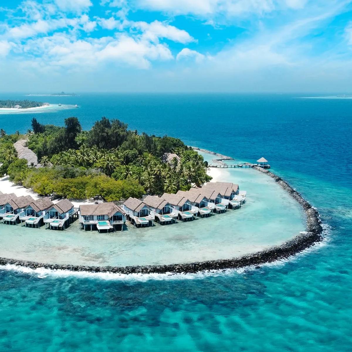 An aerial view of NH Maldives Kuda Rah Resort's small circular reef island, ringed by a rock breakwater with overwater villas stepping into the lagoon