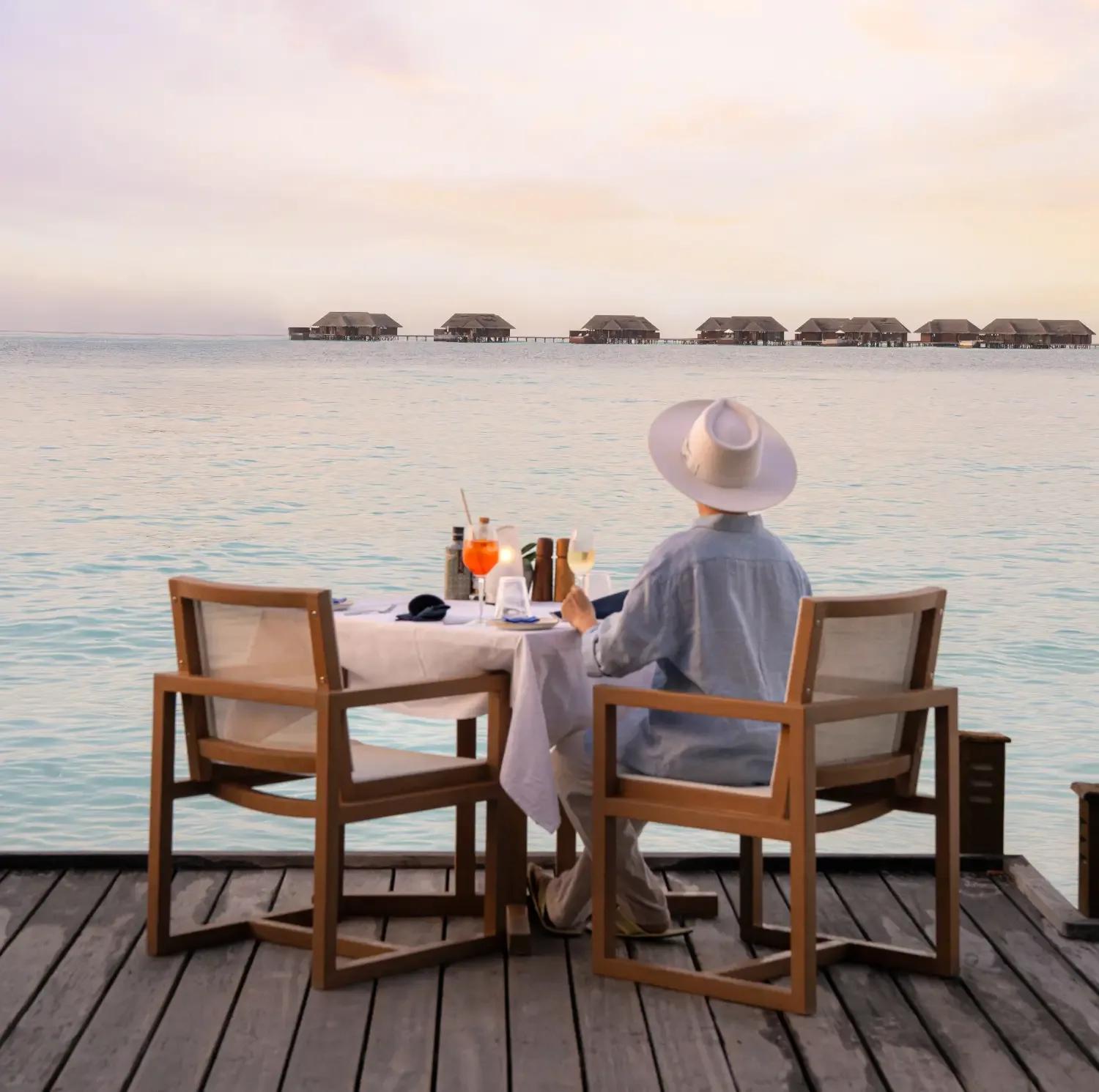 A solo diner at a lagoon-side table at Vilu Restaurant & Bar, overwater villas visible across the water at Conrad Maldives Rangali