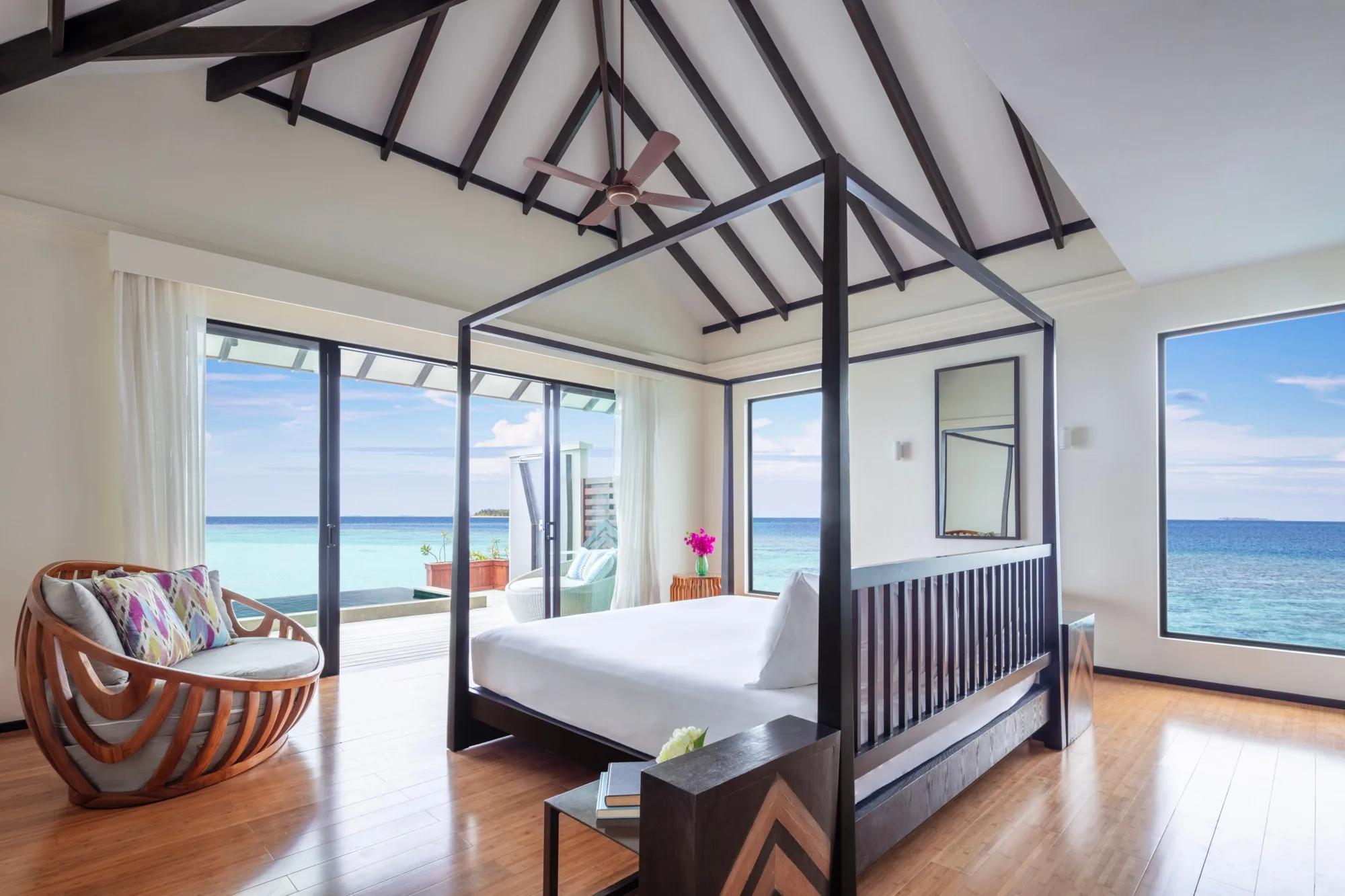 An Over Water Pool Suite's four-poster bed beneath vaulted timber beams, open sea visible through floor-to-ceiling windows on both sides and a private pool deck through the sliding doors