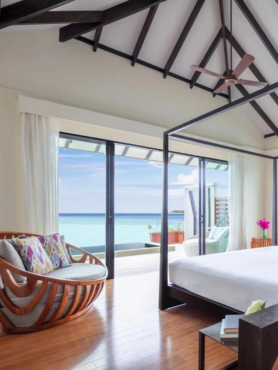 An Over Water Pool Suite's four-poster bed beneath vaulted timber beams, open sea visible through floor-to-ceiling windows on both sides and a private pool deck through the sliding doors