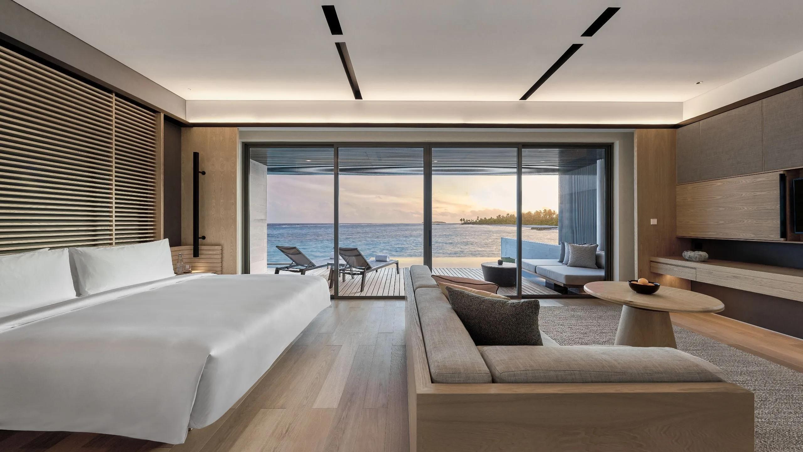 A Sunset Overwater Pool Villa's bedroom lit by the setting sun through floor-to-ceiling glass