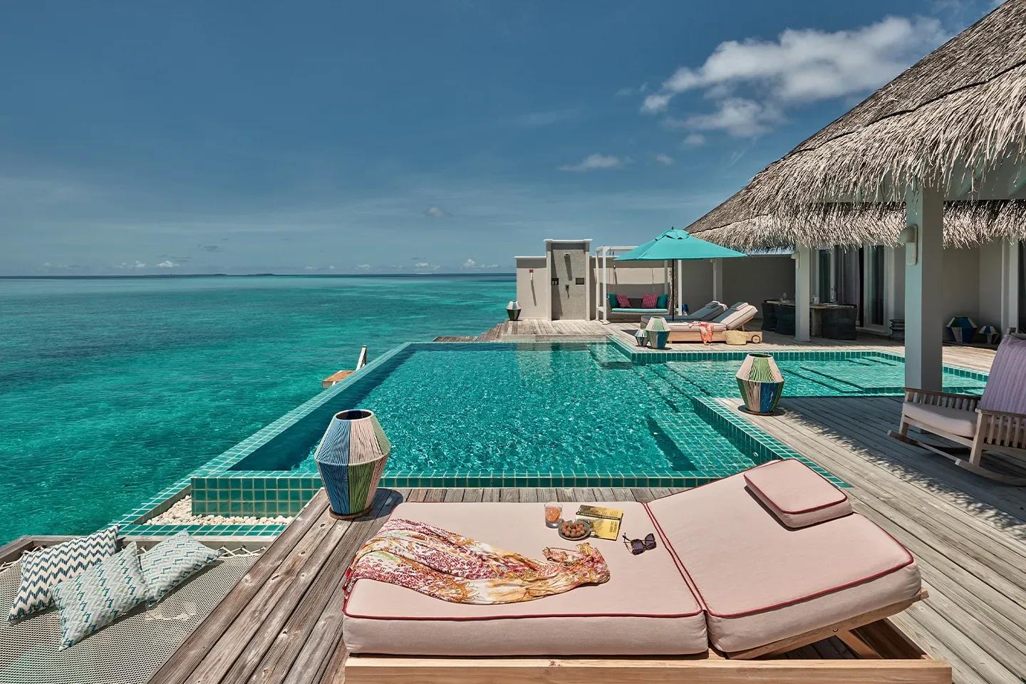 A Two Bedroom Rockstar Villa's infinity pool deck at midday, a cabana swing and striped umbrella beside the thatched roof, sun loungers set at the water's edge