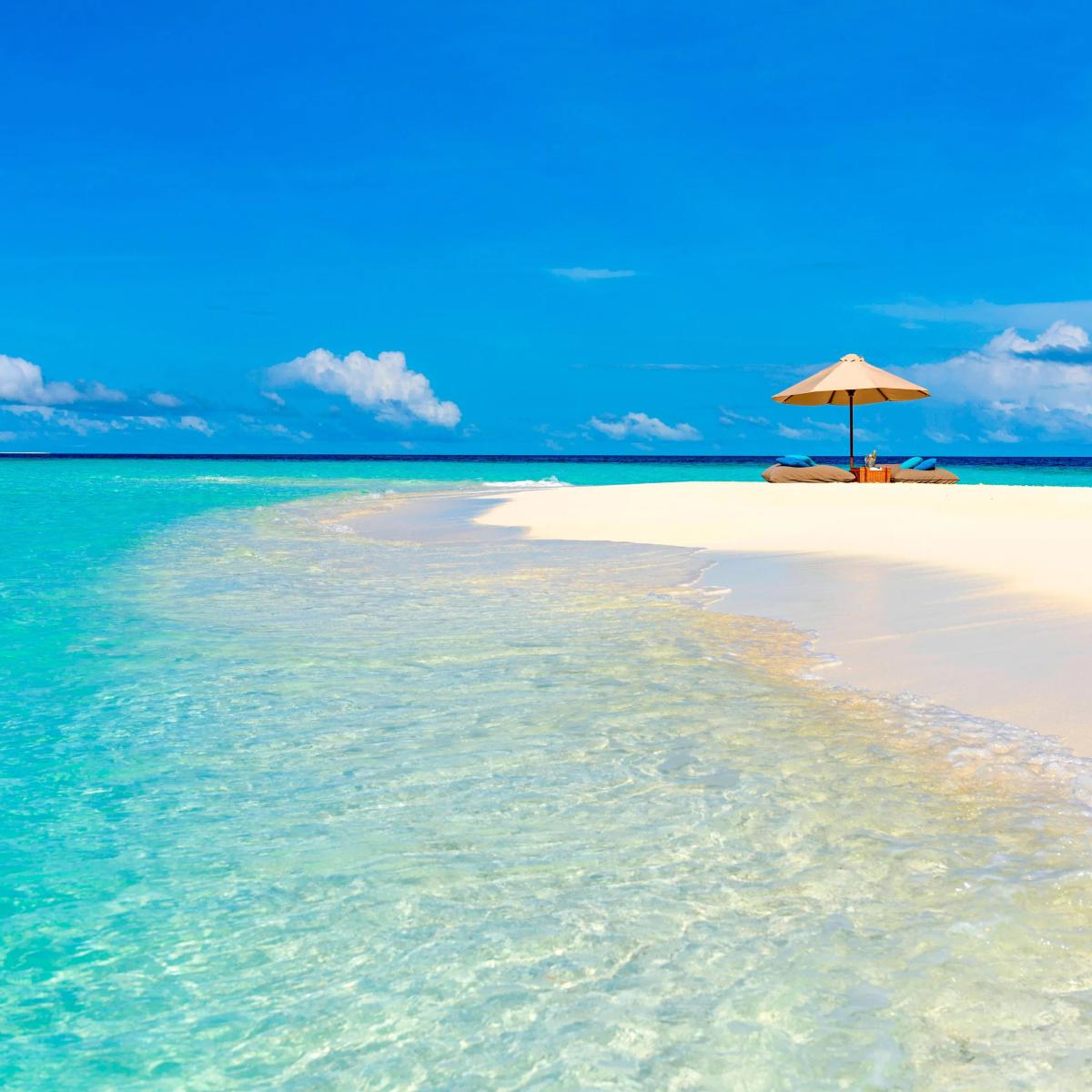 A shaded picnic setup with cushions and an umbrella on a sandbank, with clear water on both sides