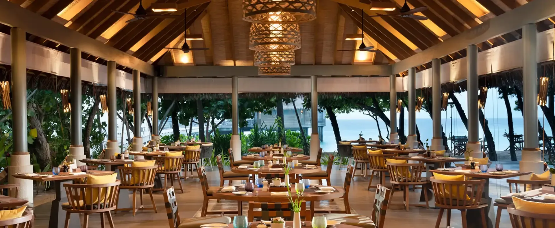 Amaany's open-air dining pavilion set beneath a high thatched roof, tables laid for dinner with the overwater villas and lagoon visible beyond