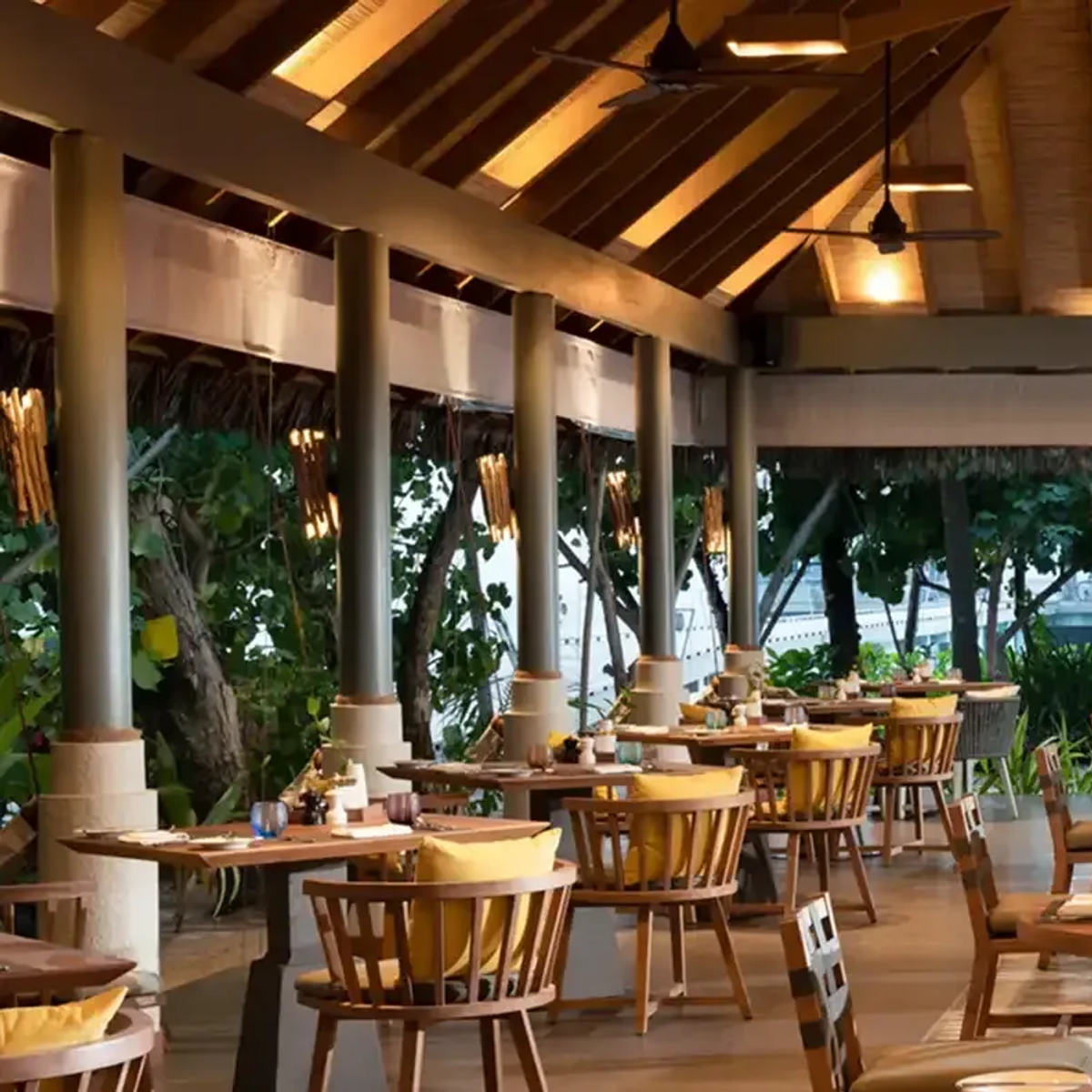 Amaany's open-air dining pavilion set beneath a high thatched roof, tables laid for dinner with the overwater villas and lagoon visible beyond