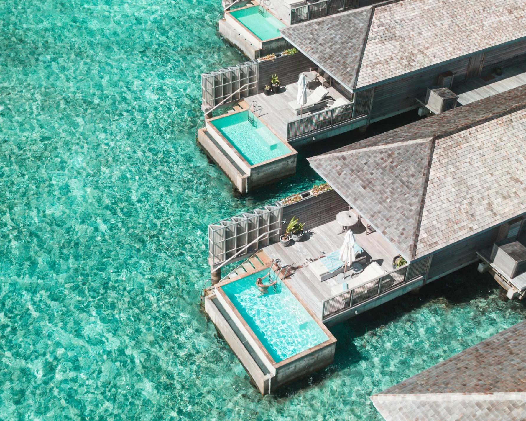 A close aerial view of two overwater villa pools cantilevered over clear turquoise water, a guest visible in one plunge pool