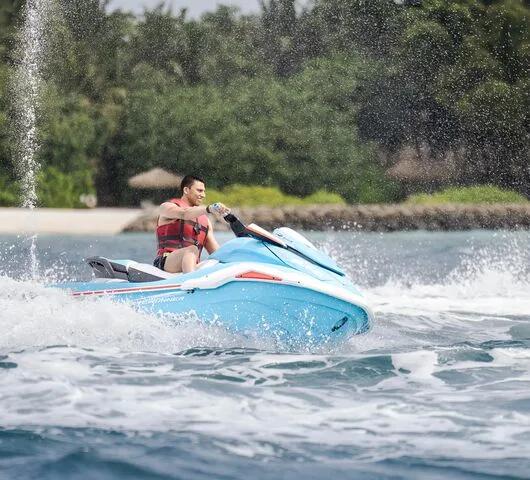 A guest in a red life vest riding a jet ski, spray fanning out behind, with the island's tree line in the background