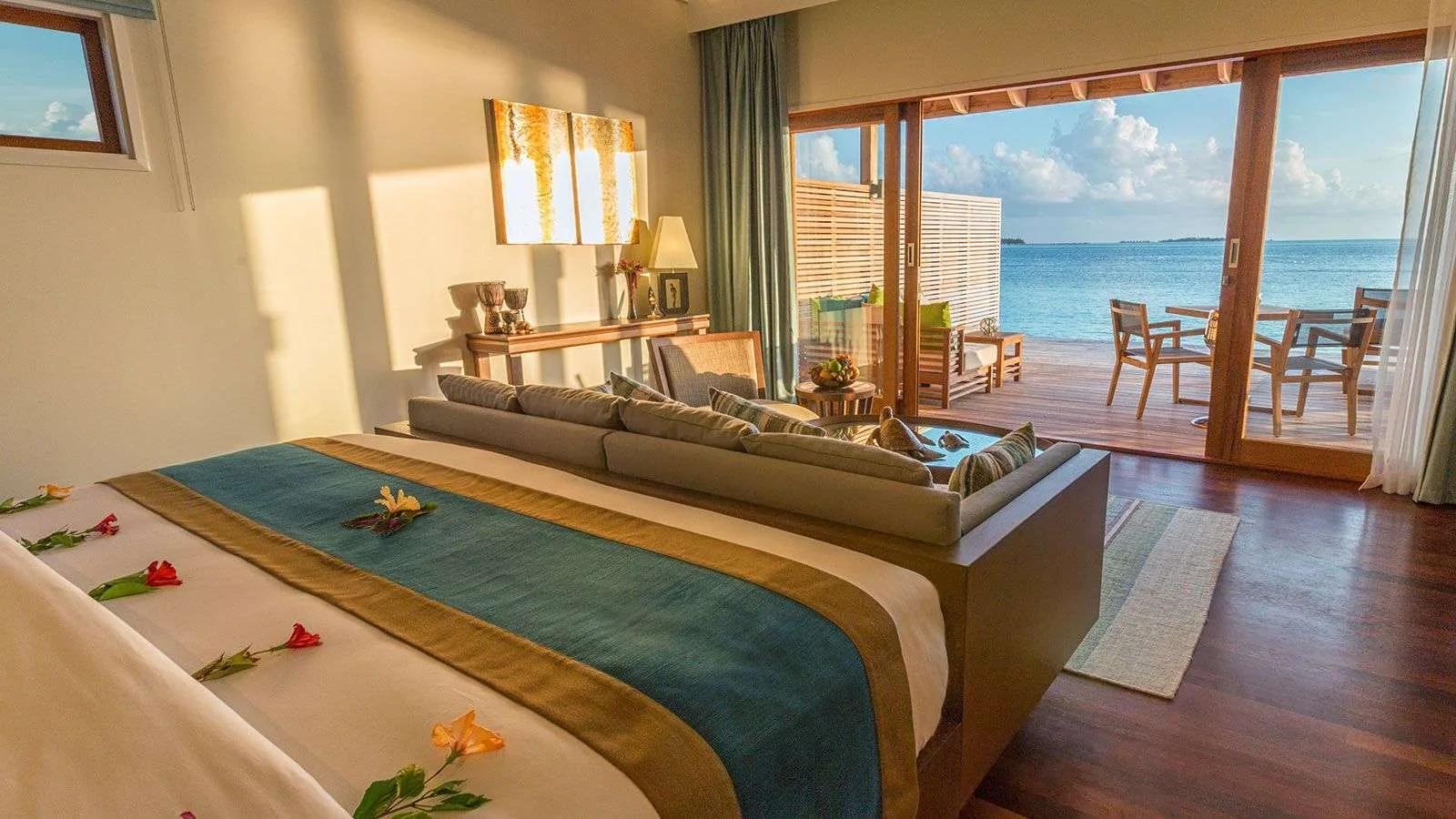 An Ocean Villa's bedroom opening onto its overwater deck, with a dining table set for two above the lagoon