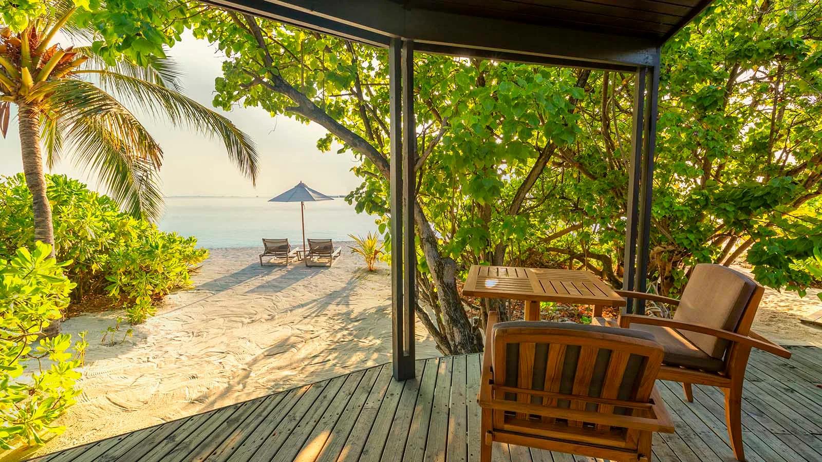 A Jacuzzi Beach Villa's shaded porch looking out through palm trees to sun loungers on the beach