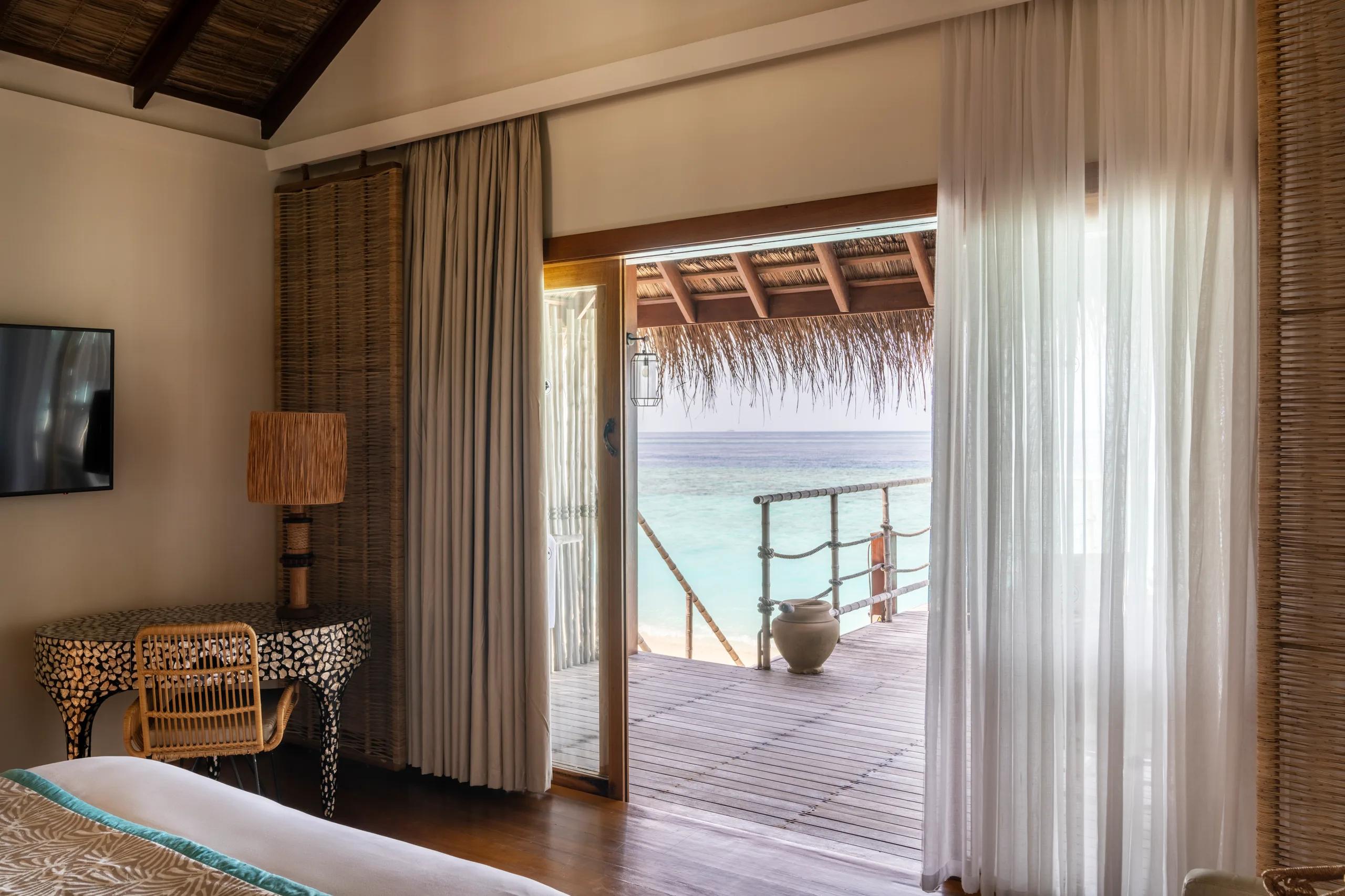 Sand Villa bedroom with sheer curtains drawn back to a thatched-roof terrace and the lagoon beyond