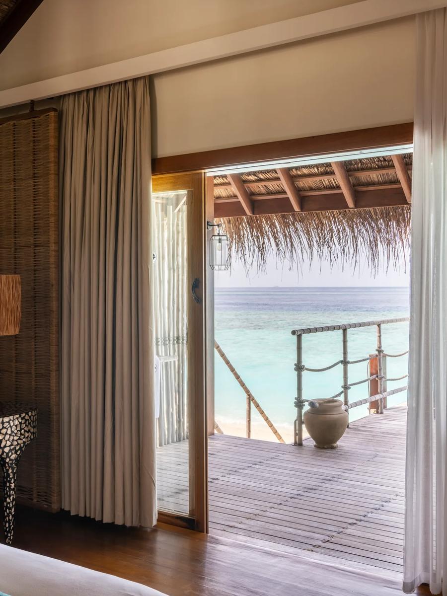 Sand Villa bedroom with sheer curtains drawn back to a thatched-roof terrace and the lagoon beyond
