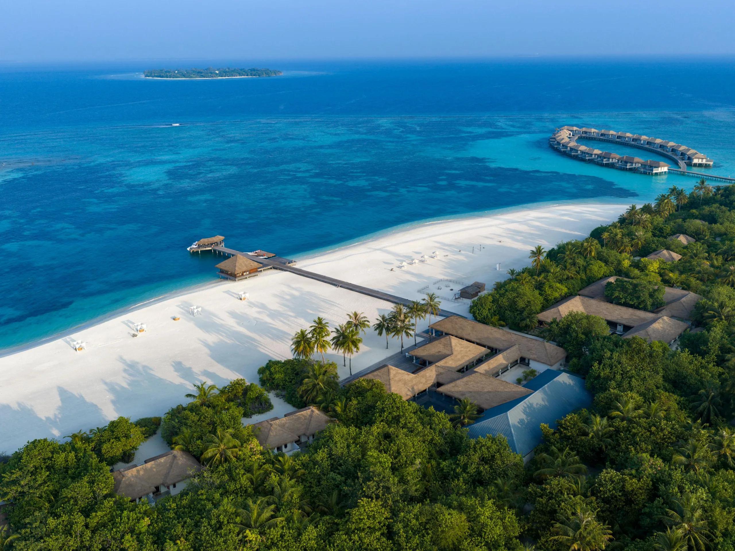 Wide aerial of Noku Maldives in late-afternoon light, beach villas lining the shore beside the overwater villa jetty