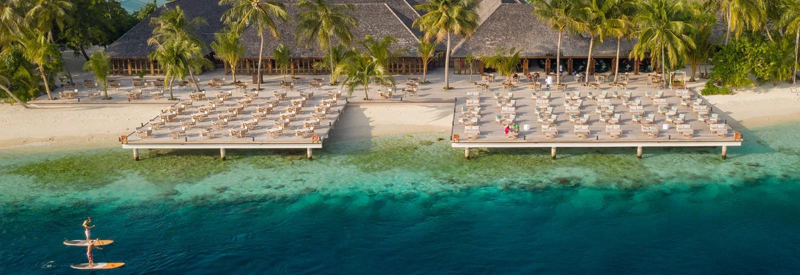 Aerial view of Vilamendhoo's twin overwater sun-deck platforms above the house reef, with a paddleboarder crossing the lagoon