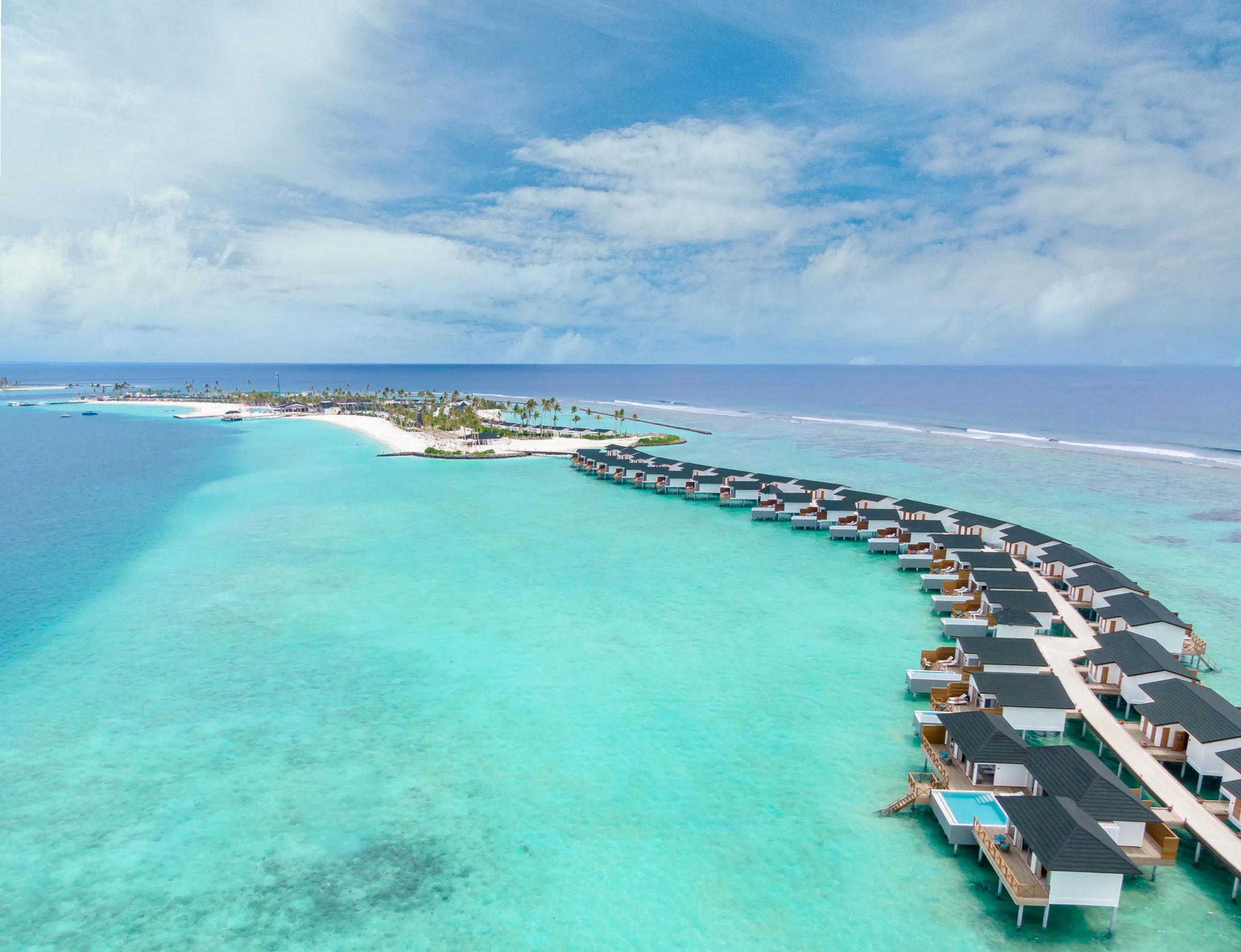A straight-on aerial of Joy Island's full row of overwater Lagoon villas reaching out from the beach into a graded turquoise lagoon