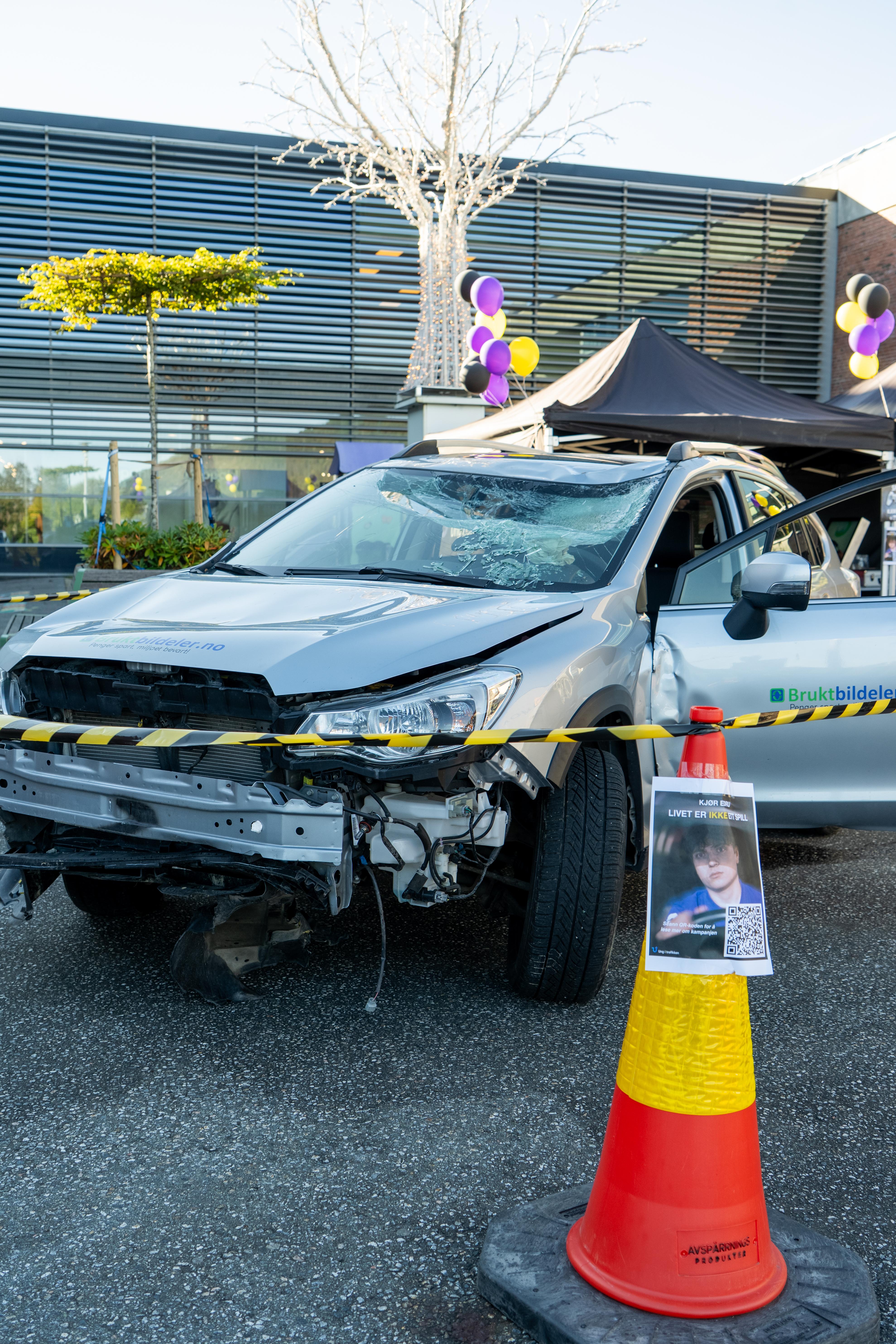 En stor kjegle med QR kode står foran en vrakbil som har en knust frontvindu og skadet på fronten av bilen. En sperrebånd har blitt satt opp rundt bilen.