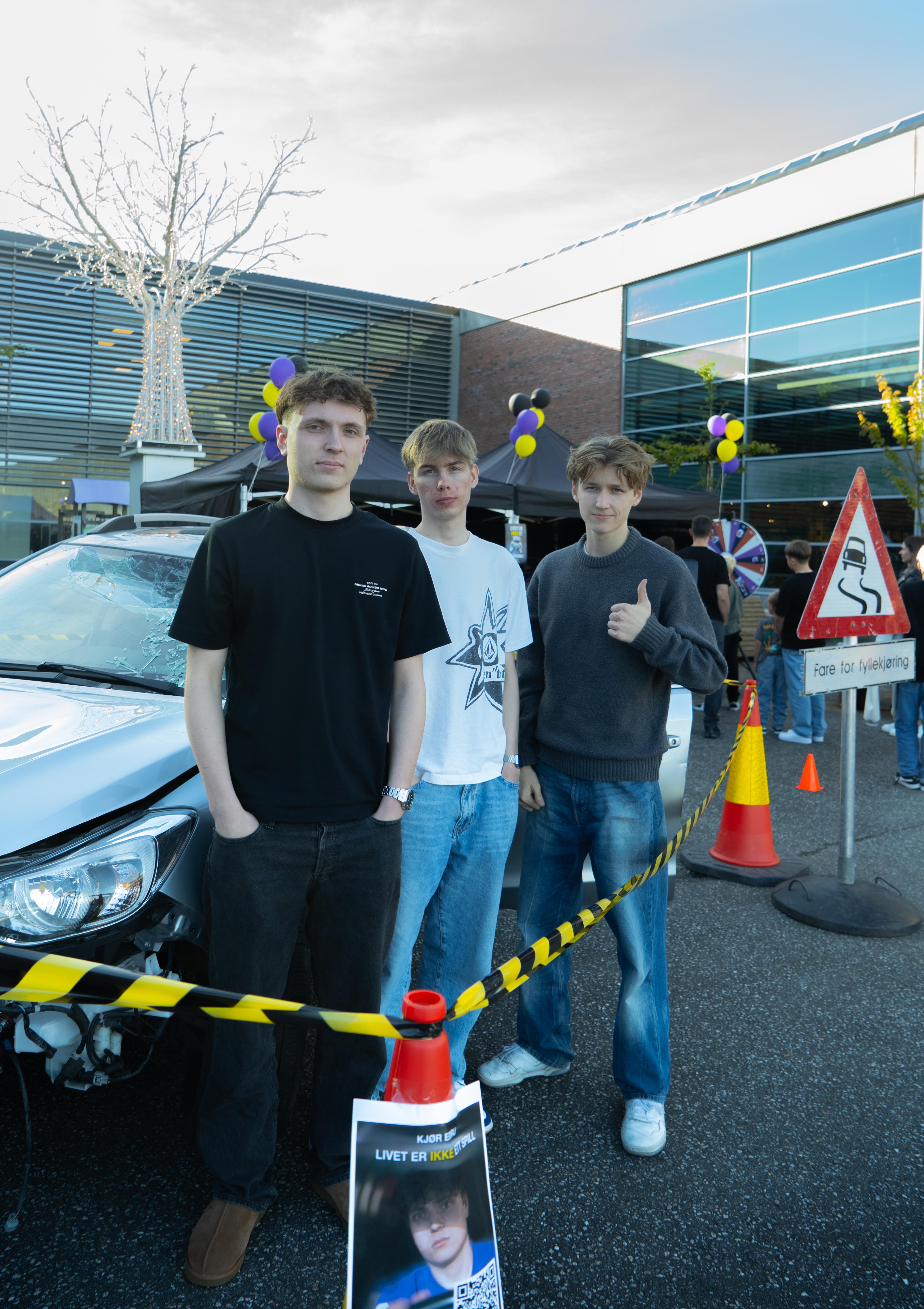 Tre av guttene bak lokalkampanjen står innenfor sperrebåndet og foran vrakbilen mens de poserer for et bilde.