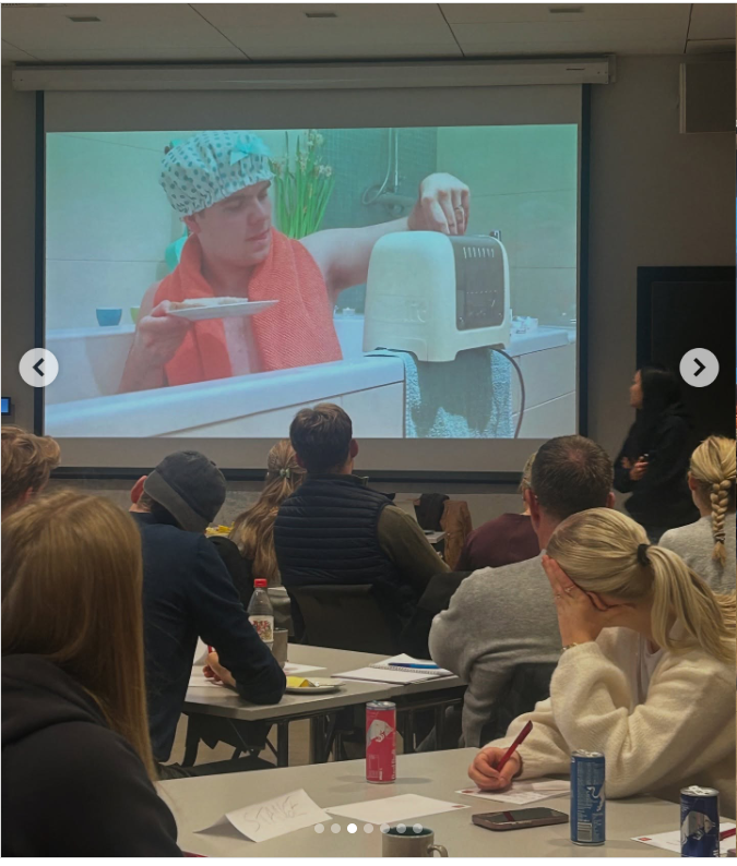 En gruppe mennesker ser på en skjerm hvor en person bader i badekar med en brødrister på siden av badekaret. 